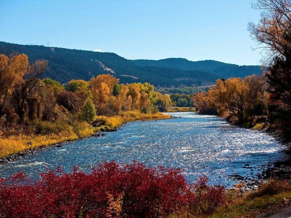 A scenic river winds through a vibrant autumn landscape, perfect for favorite day trips, surrounded by trees showcasing red, orange, and yellow foliage. The sky is clear and blue, with low hills offering a picturesque backdrop.