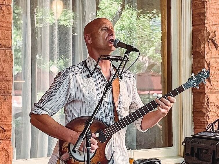 A man performs music in front of a window overlooking the courtyard, playing a guitar and using a harmonica holder. He wears a white, patterned shirt. The background features a curtain with trees visible outside, adding charm to his melodic rendition.