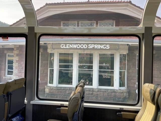 View from inside a Rocky Mountaineer train showing Glenwood Springs station. The building has a stone facade and large windows, with the train's interior seats visible in the foreground.