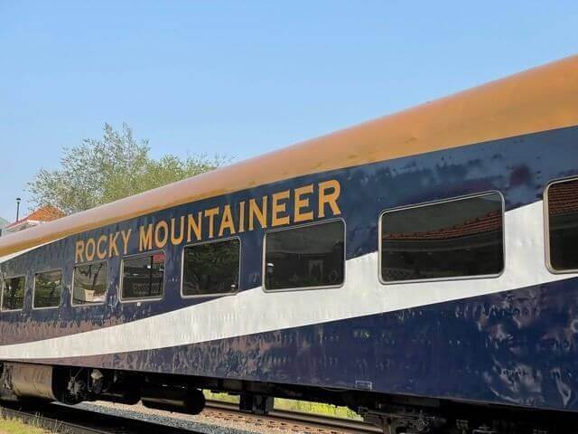 A train car proudly labeled Rocky Mountaineer glides through the landscape on a sunny day, its sleek blue and gold design accented by large windows. Under a clear sky, with just a few trees in the distance, this iconic train makes for an unforgettable journey.