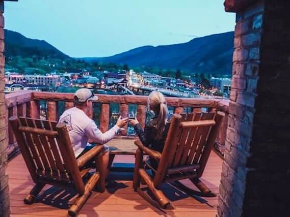 Two people sit on wooden chairs on a balcony, exuding romance as they raise their drinks overlooking a scenic view of a town nestled in a valley at dusk. Mountains loom in the background under the deepening blue sky, creating the perfect intimate setting.