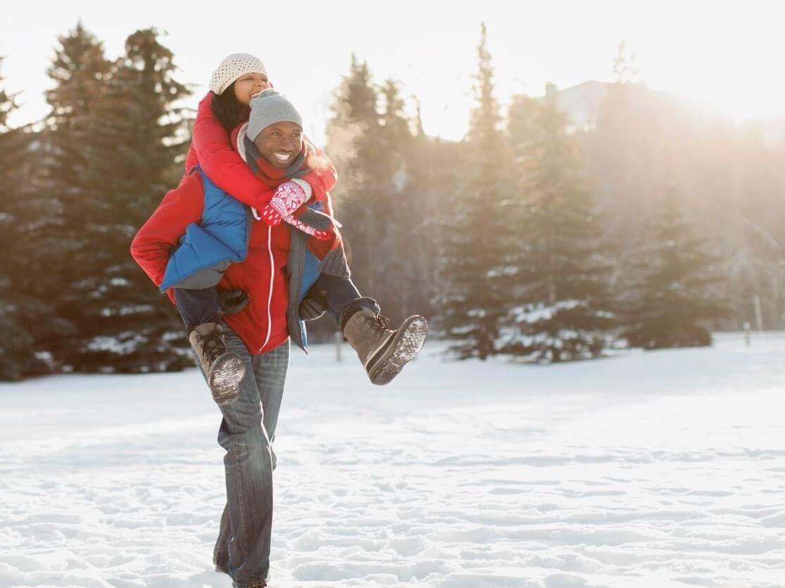 In a snowy landscape dotted with evergreen trees, a person romantically carries another on their back. Both are bundled in winter clothing, complete with hats and gloves. Sunlight filters through the trees, casting a warm glow on the scene, adding to the enchanting atmosphere.