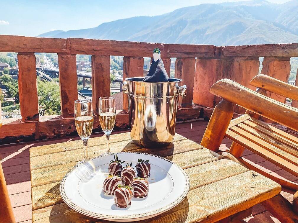 A wooden balcony with two chairs overlooks a romantic view of the mountains. A table holds a plate of chocolate-covered strawberries, two glasses of champagne, and a bottle chilling in an ice bucket.