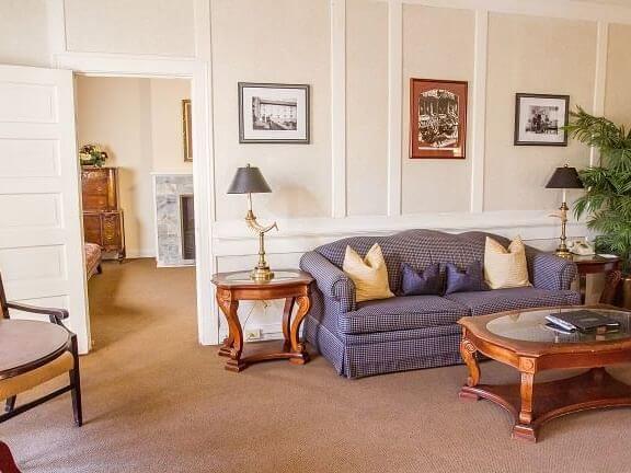 A cozy living room embodies the suite life with a checkered sofa and yellow pillows, flanked by wooden side tables with lamps. Artwork adorns the walls, while an open door offers a glimpse of the bedroom's inviting chest of drawers and charming fireplace.