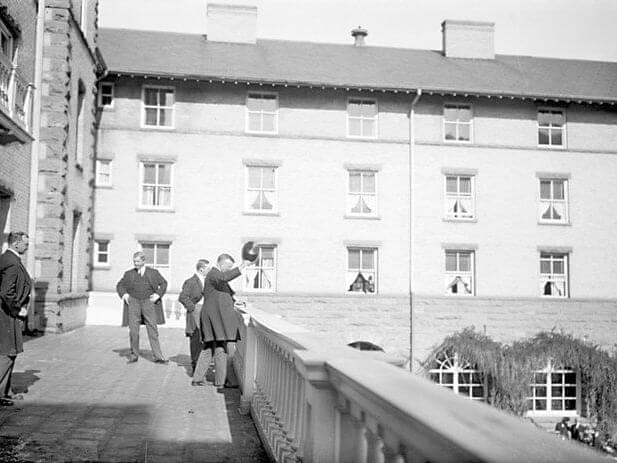A black and white photo captures several men in suits on a linked balcony or terrace, set against a large brick building. One man appears to be waving his hat. The building features multiple windows, with some curtains drawn, adding an air of mystery to the scene.