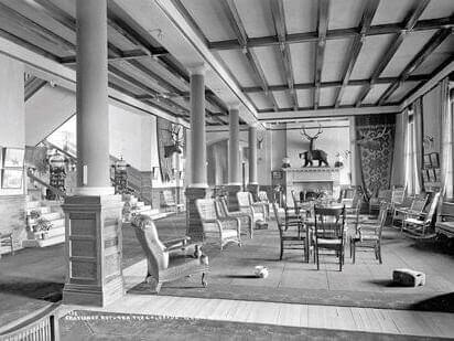 The vintage hotel lobby exudes charm with its high ceiling, large pillars, and wicker chairs. Walter's tasteful touch is evident at the heart of the room—a fireplace adorned with a mounted deer head and antlers. Sunlight streams through an entire wall of windows, casting a warm glow across the inviting space.