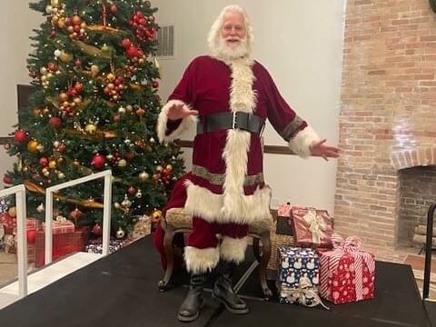A person dressed as Santa Claus stands cheerfully beside a decorated Christmas tree and several wrapped gifts, embodying beloved holiday traditions. The room has a cozy ambiance with a brick fireplace in the background.