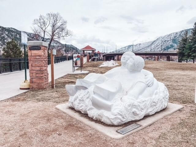 A white sculpture of two figures reclining together is displayed in a park setting, offering perfect photo opportunities. A pathway lined with trees and park signs leads to a bridge with mountains in the background under a cloudy sky.