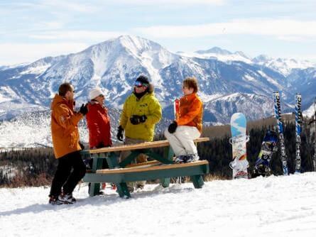 A group of four people in winter clothing stands and sits on a picnic table, basking in the sunlight across a snowy landscape. Snowboards are propped against the table, with majestic mountains visible under a clear blue sky.