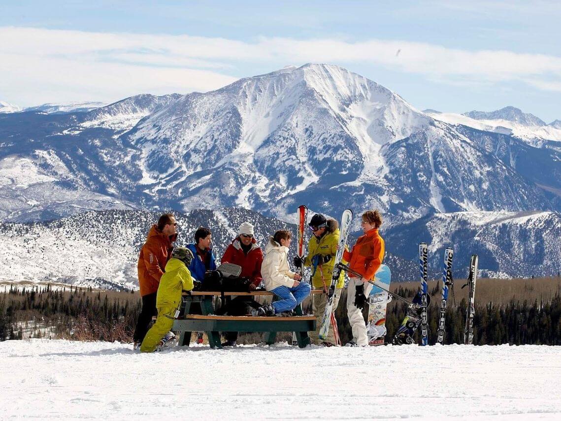 A group of people in colorful ski gear gather around a picnic table on the snowy mountain, enjoying the great outdoors. Skis and snowboards rest upright in the snow, while majestic mountain peaks rise against a clear blue sky.