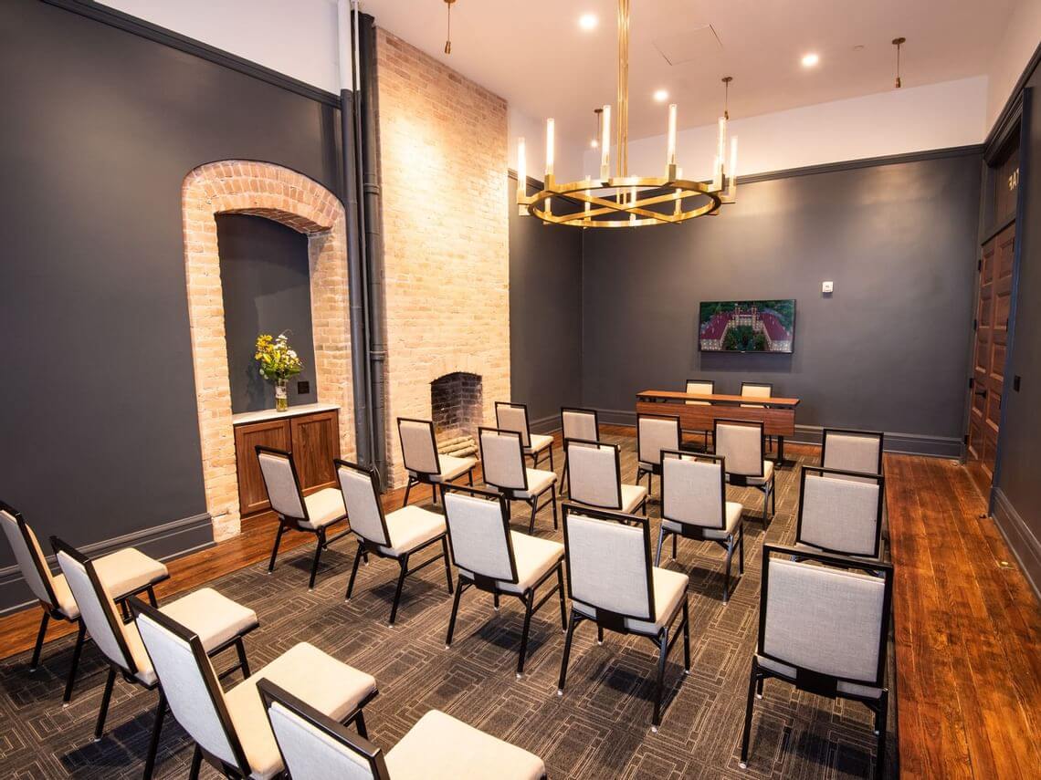 A modern, well-lit meeting space features rows of beige chairs facing a wooden table and a mounted TV. The room has dark walls, a brick accent, and a large circular chandelier hanging from the ceiling. Florals decorate a side niche.