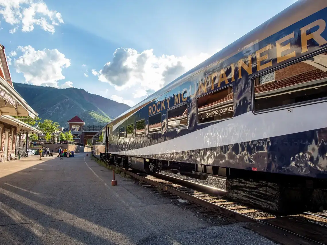 A Rocky Mountaineer train rests at a scenic platform under a clear sky, with sunlight casting playful shadows. Majestic mountains and a vintage-style building provide a picturesque backdrop, completing this charming tableau.