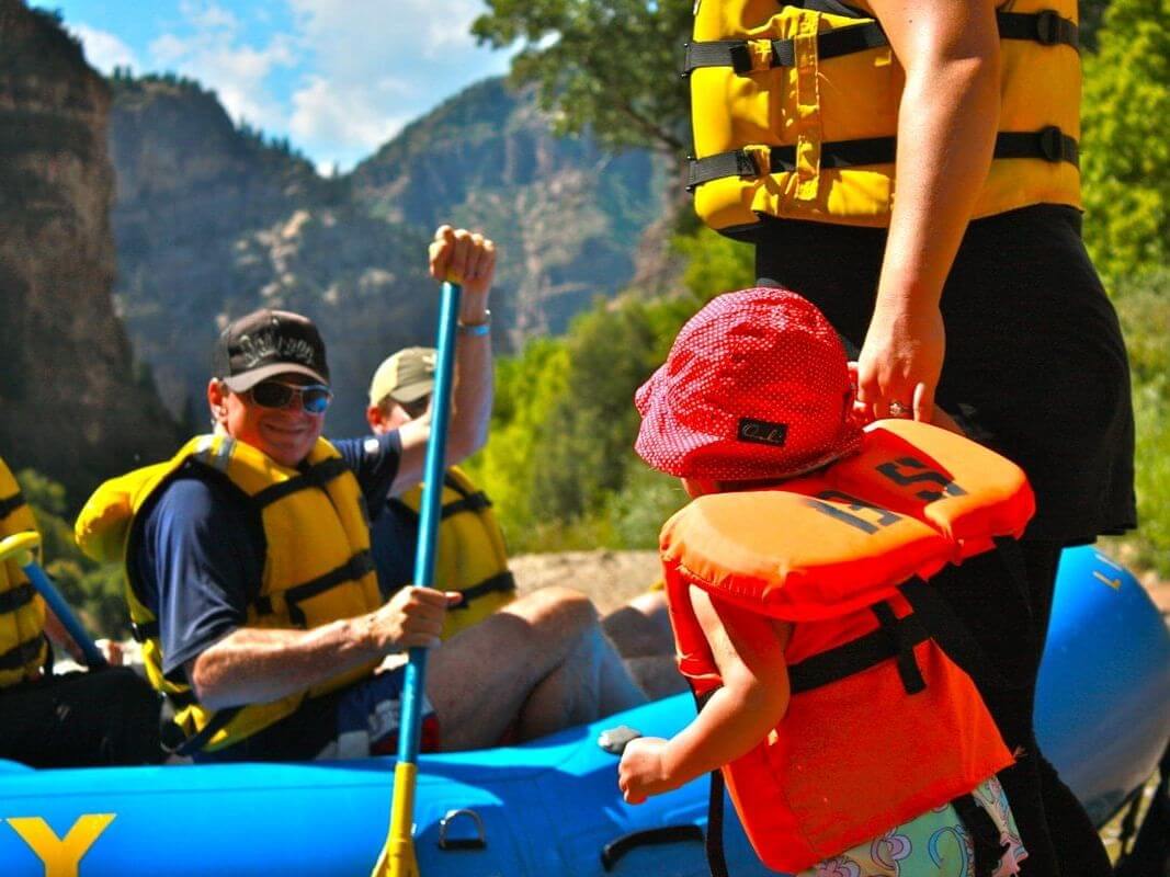 A group of people in life vests prepare for a thrilling rafting adventure, a memorable event to check off their vacation bucket list. A child wearing an orange life vest and red sun hat stands in the foreground. Trees and mountains provide a stunning backdrop.
