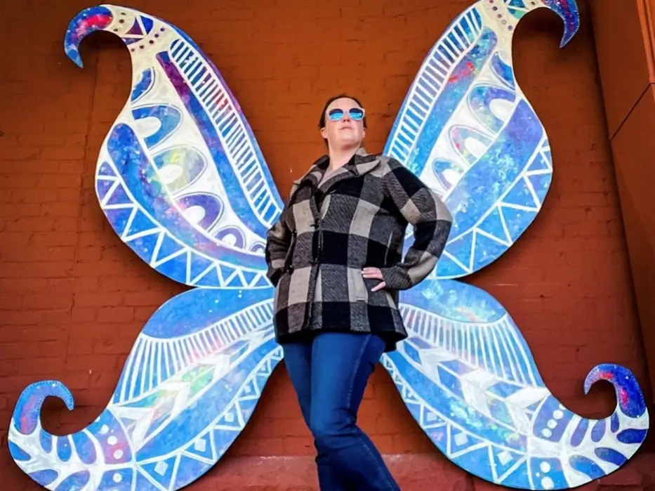Clad in a black and white checkered jacket and jeans, a person stands confidently in front of vibrant butterfly wings painted on a wall—a perfect photo opportunity. Sporting blue sunglasses, they pose with their hands tucked casually into their jacket pockets.