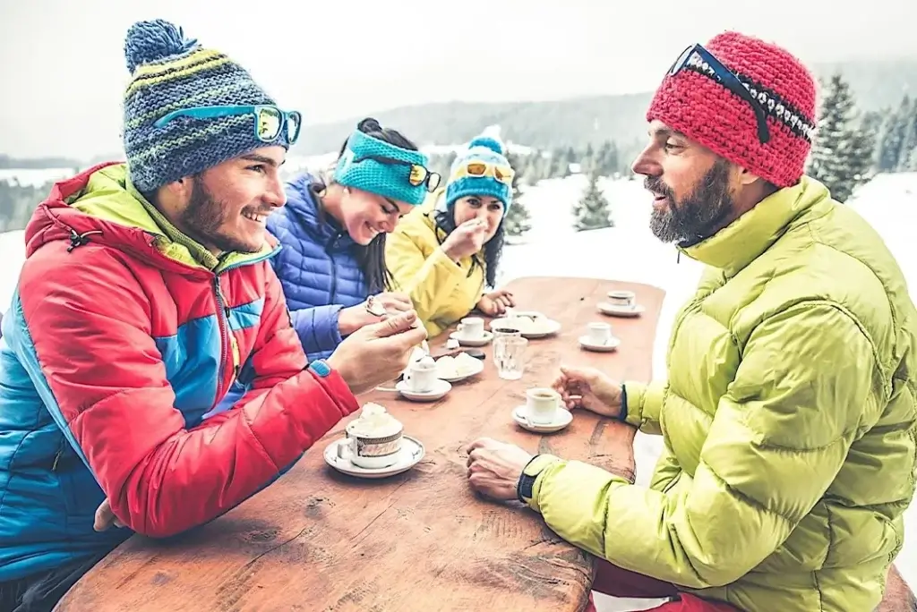 Four people in winter gear sit at a wooden table outdoors, enjoying hot drinks