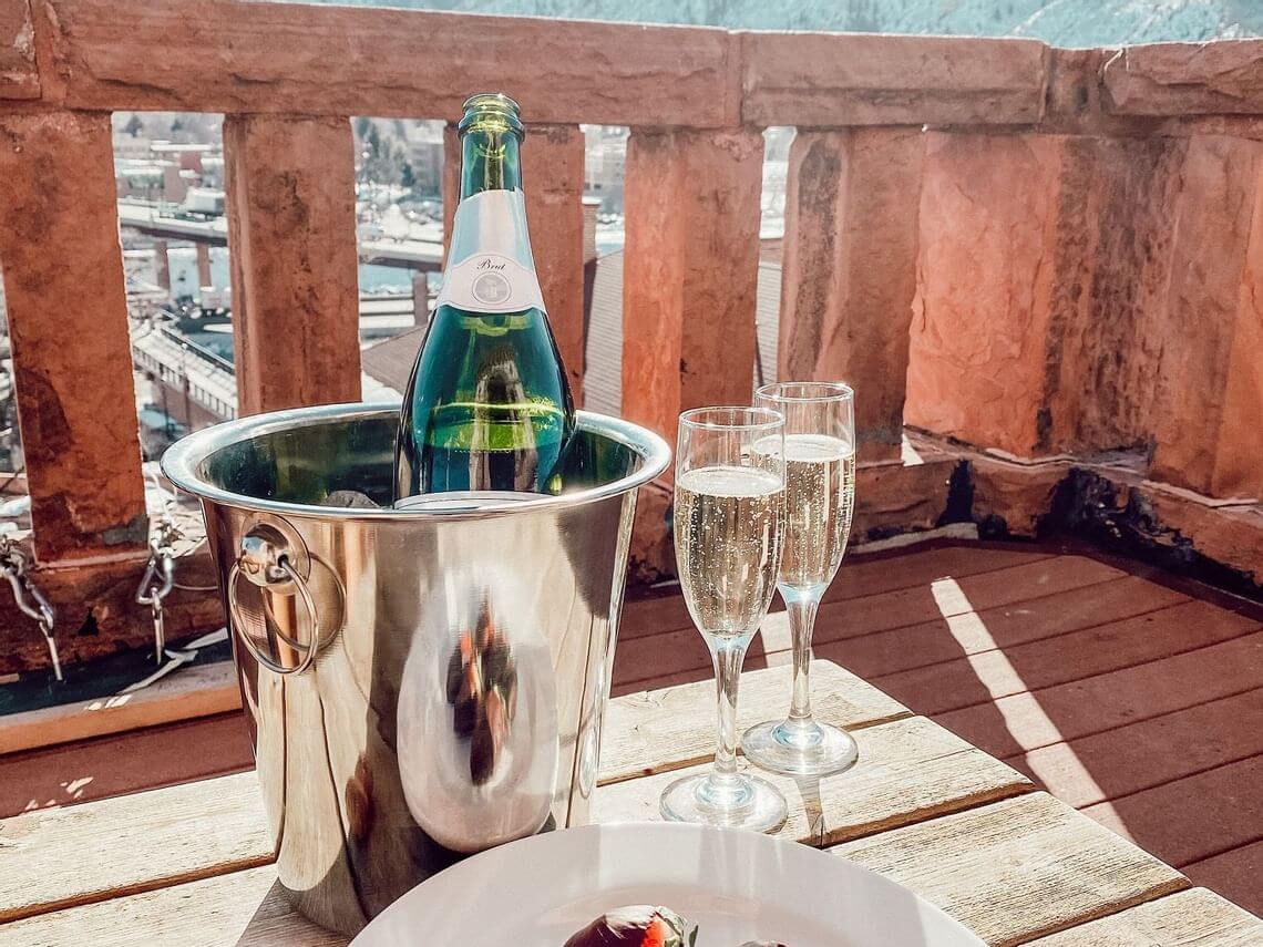 A bottle of sparkling wine sits in an ice bucket with two filled glasses on a wooden balcony, perfect for a romantic getaway. Sunlight dances on the rustic railing as you soak in the scenic view.