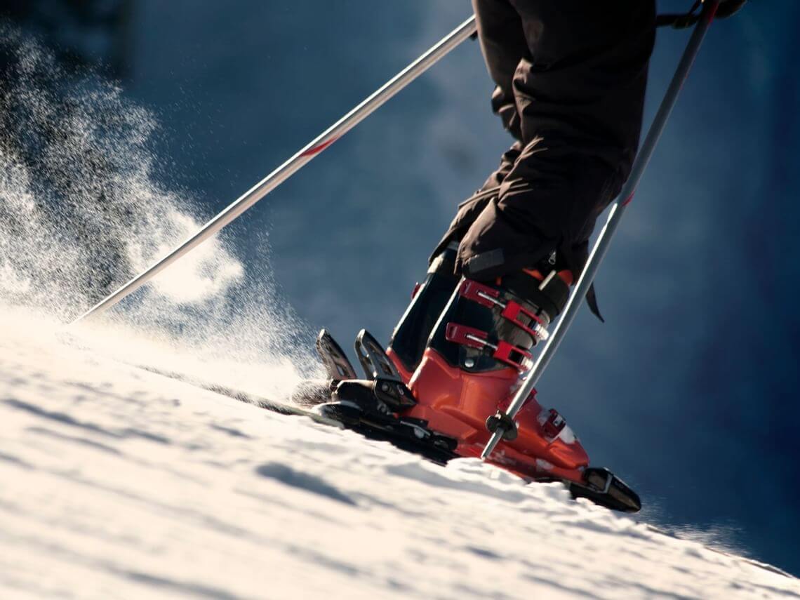 A skier wearing bright orange ski boots and holding poles glides smoothly down a snowy slope, kicking up powder under a clear blue sky, as if ready for the X Games.