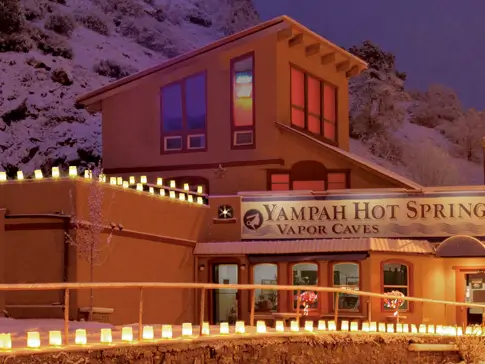 A warmly lit building covered in snow displays a sign reading Yampah Hot Springs Vapor Caves. The structure is adorned with glowing lanterns, and the surrounding winter landscape is gently snow-covered, creating a cozy nighttime scene.
