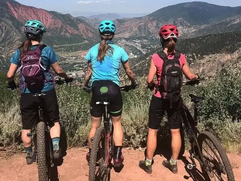 Three female cyclists in helmets and gear stand with their bikes, enjoying the view of Glenwood Springs below