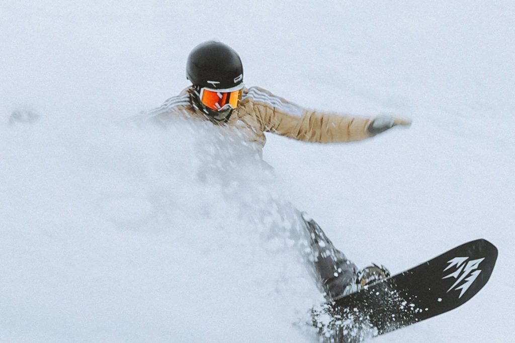 A snowboarder dressed in a beige jacket, black helmet, and reflective goggles carving through fresh snow