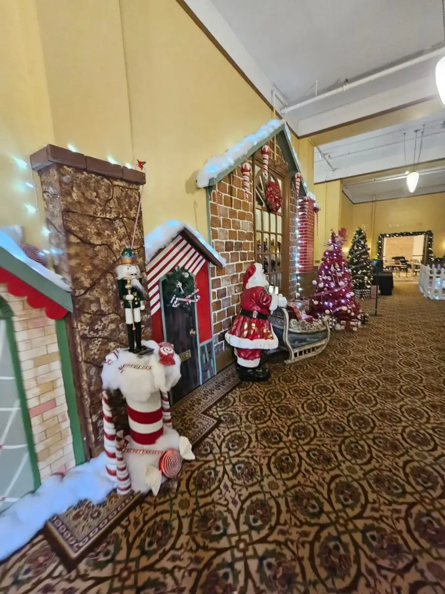 A festive hallway bursts with Christmas cheer, featuring toy soldiers, candy canes, and a Santa Claus figure with a sleigh. Christmas trees twinkle brightly as walls resembling gingerbread houses envelop the scene, while an ornate patterned carpet completes the whimsical atmosphere.