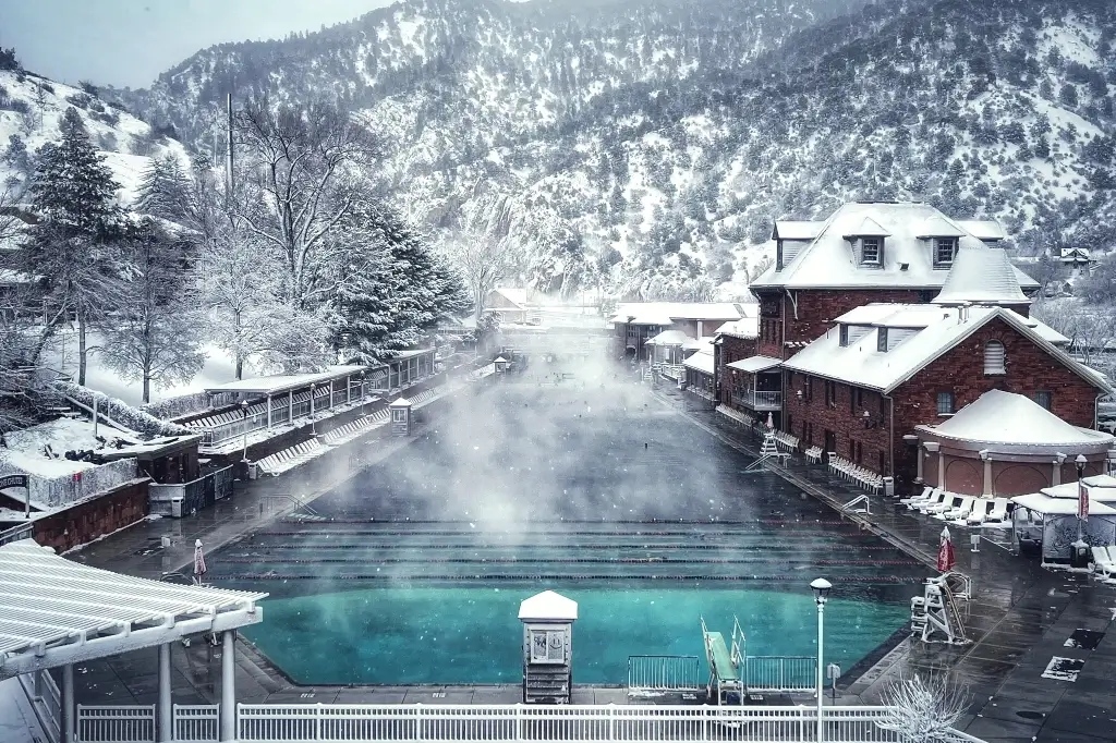 The snow-covered landscape features a large, steaming outdoor pool, perfect after a long day of skiing. Surrounded by buildings and trees, mountains in the background create a wintery atmosphere. A serene smaller pool in the foreground rests under a blanket of snow.