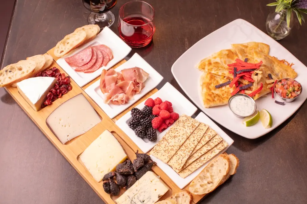 A wooden platter showcases assorted cheeses, salami, and prosciutto alongside 72 hours-aged crackers, bread, berries, and dried fruits.