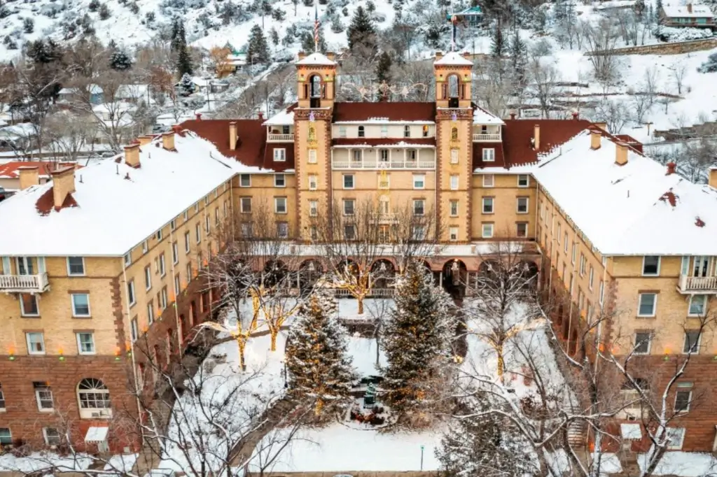 Aerial view of a historic ski hotel surrounded by snow. The U-shaped building, with two towers and a red roof, nestles among snow-covered trees and pathways in the courtyard. The background showcases a wintery landscape with hills and trees, perfect for ski enthusiasts.