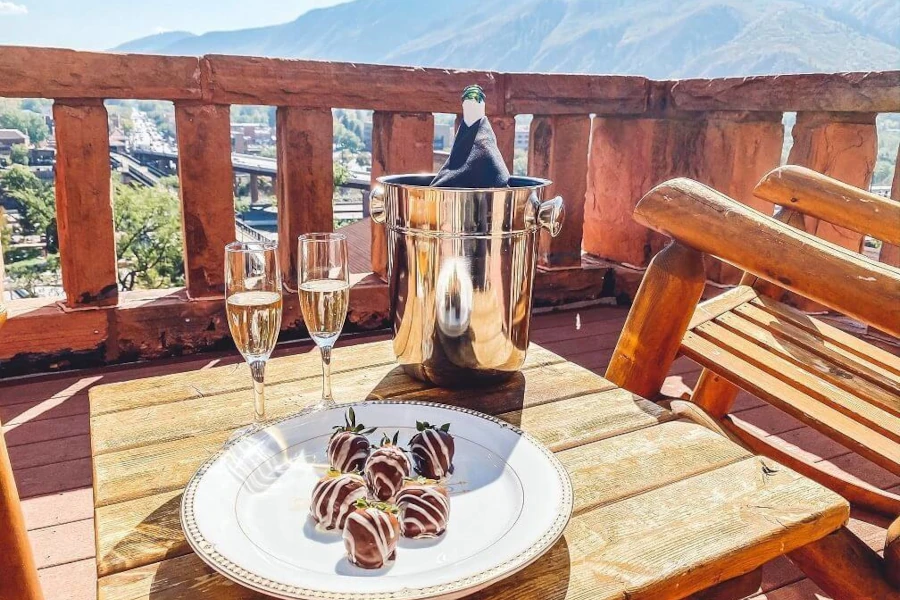 A wooden patio set, with a touch of romance, showcases two glasses of sparkling wine and a chilled bottle in an ice bucket. A white plate holds strawberries dipped in chocolate. In the background, the majestic Rockies frame a scenic valley view under a clear sky.