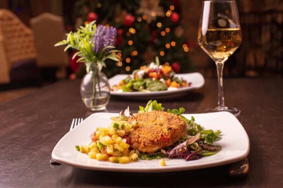 A gourmet dish of a breaded patty sits on a bed of mixed greens, garnished with diced vegetables. A glass of white wine complements the romantic setting. In the background, another dish and a small vase with flowers are visible, with festive lights blurred like a cozy hideaway in the Rockies.
