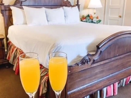 A cozy bedroom exuding romance, with a large wooden bed draped in white linens and several pillows. In the foreground, two glasses of orange juice await on a surface. A lamp and flower arrangement sit on a bedside table, creating an inviting retreat reminiscent of the Rockies.