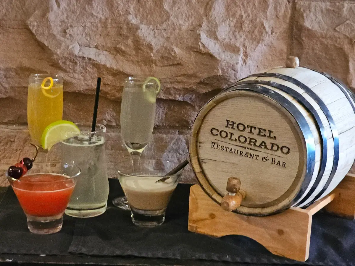 A small wooden barrel labeled Hotel Colorado Restaurant & Bar sits on a table next to five cocktails.