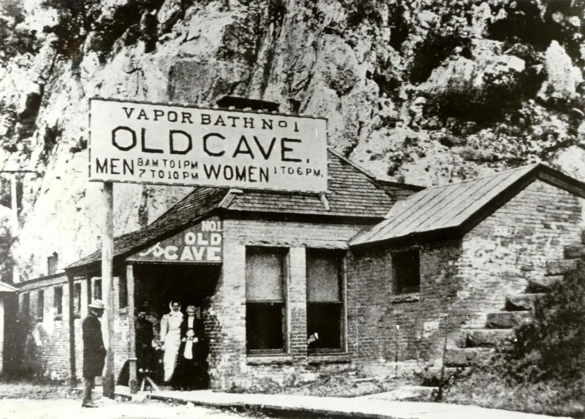 A black-and-white photograph captures a rustic building nestled against a rocky hillside. A sign reads Vapor Bath No. 1 Old Cave, reminiscent of the Yampah Spas Vapor Caves, with hours for men and women. Several people gather near the entrance, while a stone staircase ascends to the right.