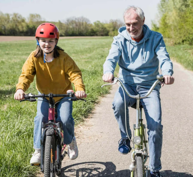 An older man and his grandkid, both smiling, ride bicycles side by side on a paved path through a green, grassy park. The girl wears a red helmet and yellow sweater; the man sports a blue hoodie on this perfect Glenwood Springs getaway.