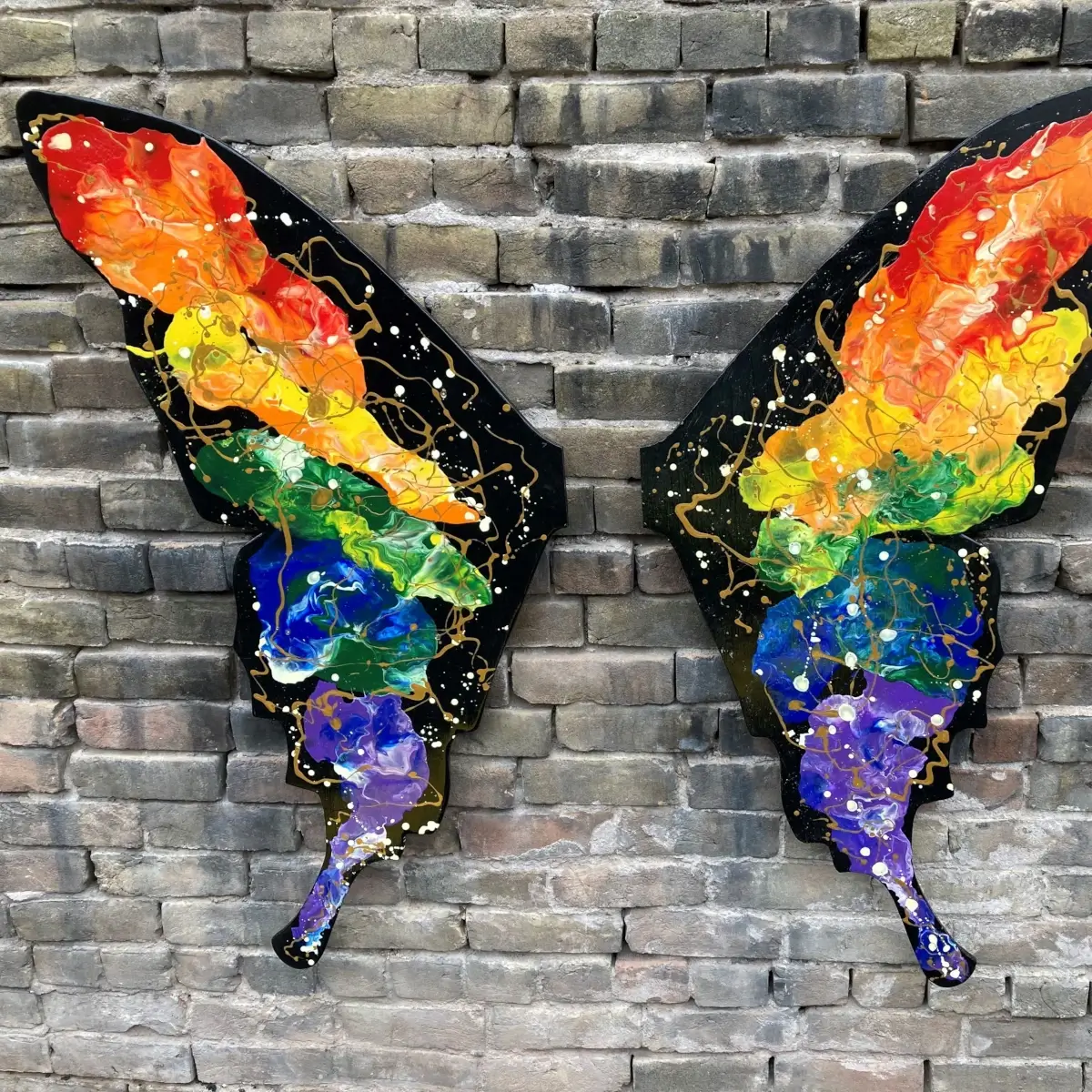 Two painted butterfly wings in rainbow colors are mounted on a brick wall, positioned so that someone standing between them appears to have wings. The wings are decorated with splashes of gold, white, and black paint.