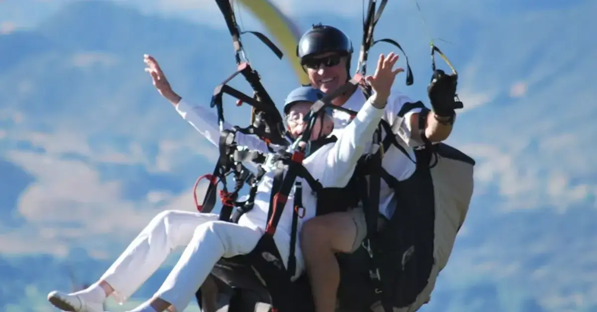 Two people paragliding in tandem, both smiling and waving, with mountains and blue sky in the background. The front person is wearing white clothes; both are wearing helmets and sunglasses.