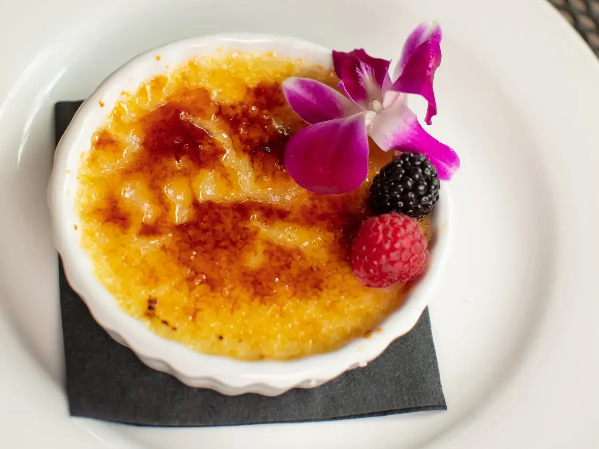A dish of crème brûlée with a caramelized sugar top, garnished with a purple orchid flower, a blackberry, and a raspberry, served in a white ramekin on a white plate.