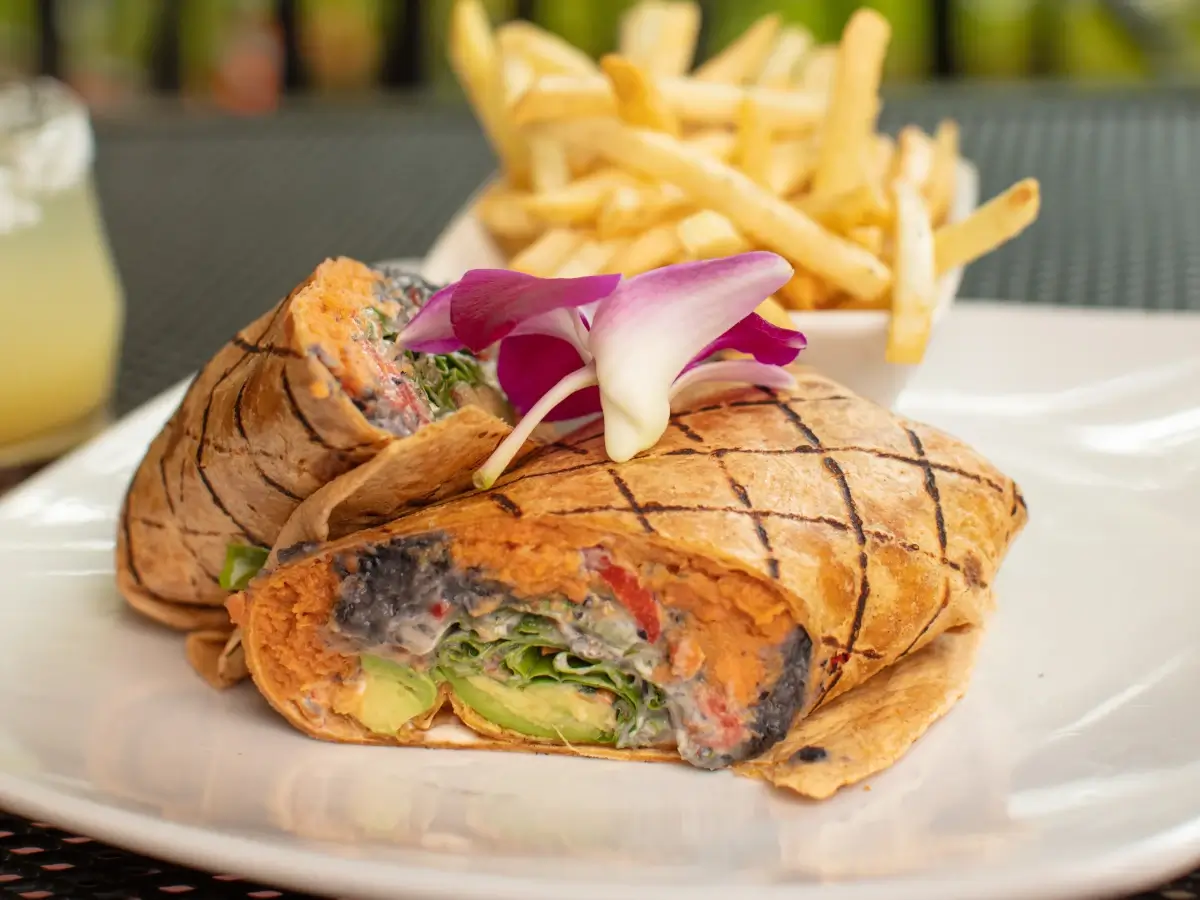 A grilled wrap filled with vegetables and black beans, cut in half and garnished with a purple edible flower, is served on a white plate with a side of French fries in the background.
