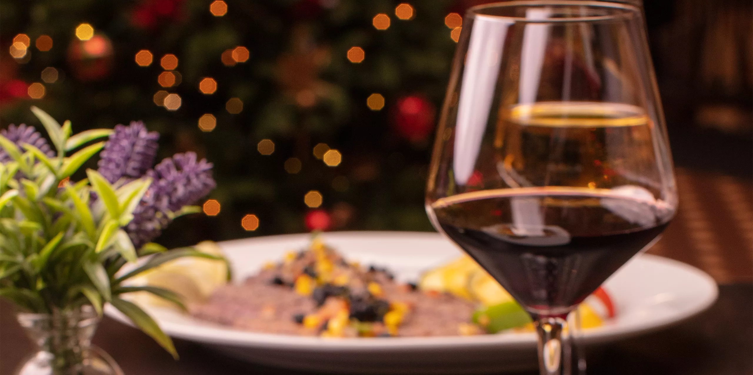 A close-up of a glass of red wine on a restaurant table, with a blurred plate of food and a small vase of purple flowers in the background. Warm, festive lights softly glow behind.