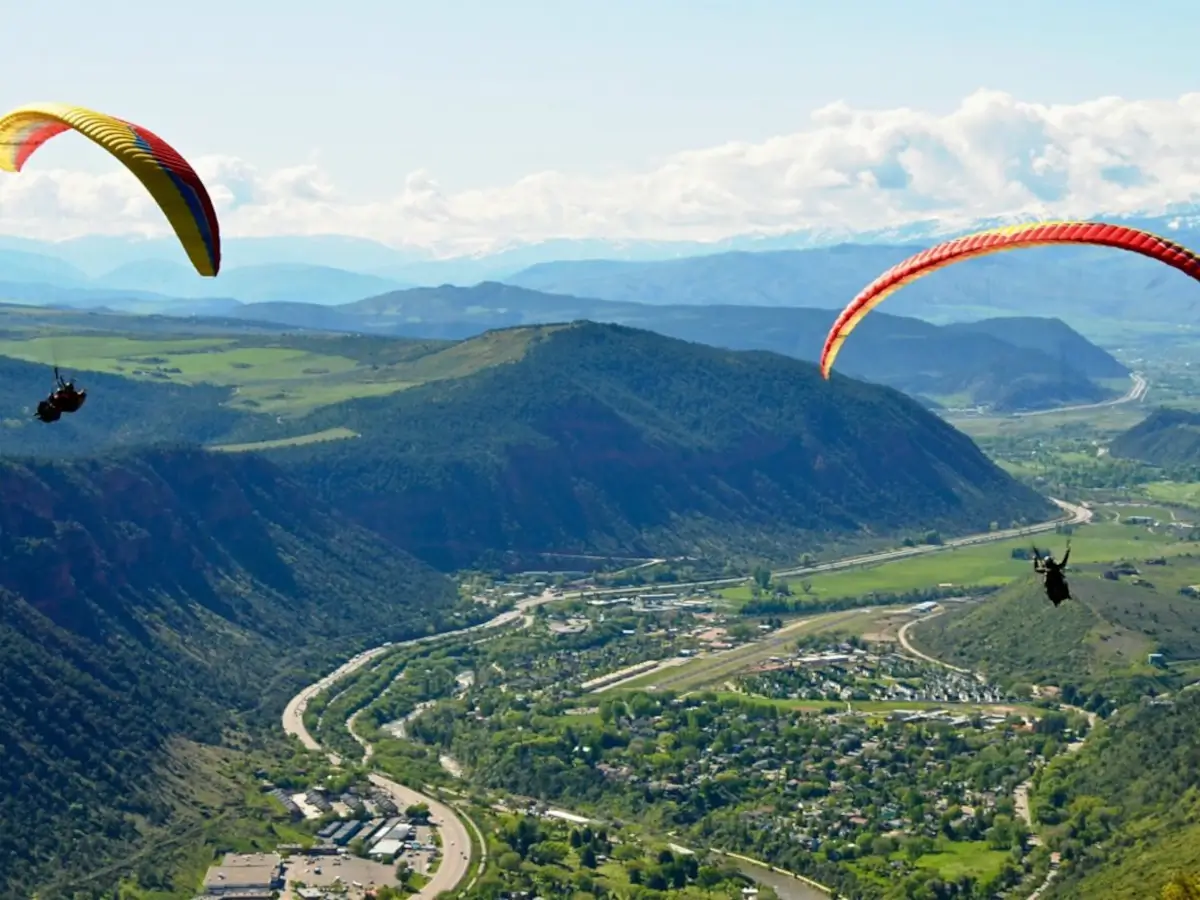 Two people paragliding high above a lush, green valley with winding roads, a small town below, and forested hills and mountains in the distance under a partly cloudy sky.
