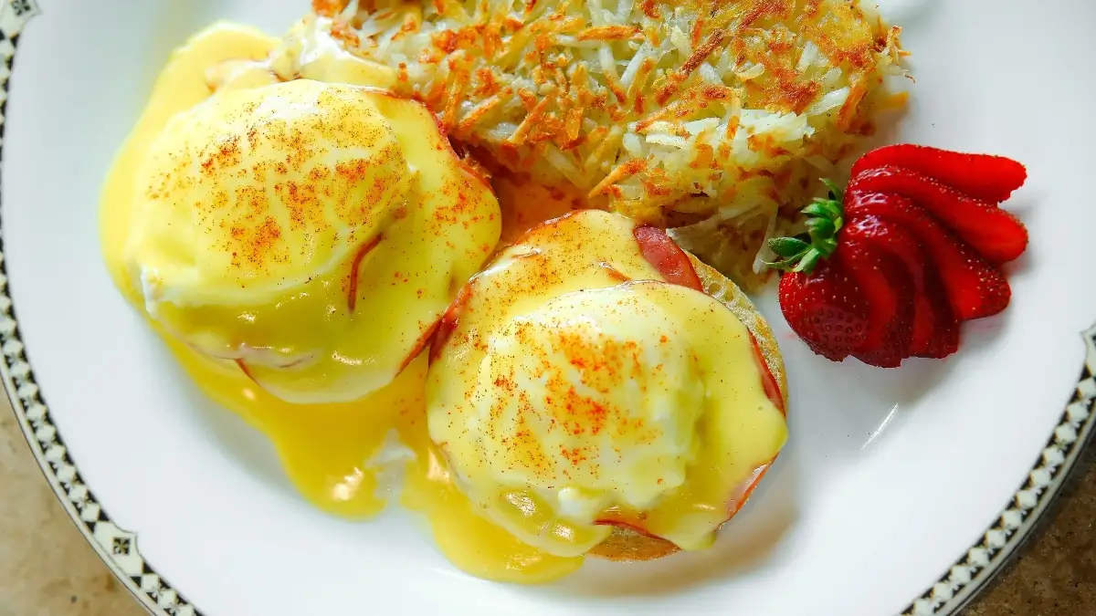A plate of eggs Benedict topped with hollandaise sauce and paprika, served with a portion of crispy hash browns and a sliced strawberry garnish.
