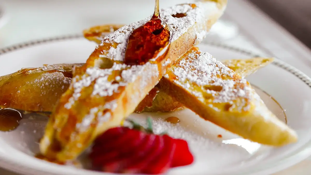 Golden slices of French toast dusted with powdered sugar, topped with a drizzle of syrup from a spoon, and garnished with a red fruit and green leaf on a white plate.