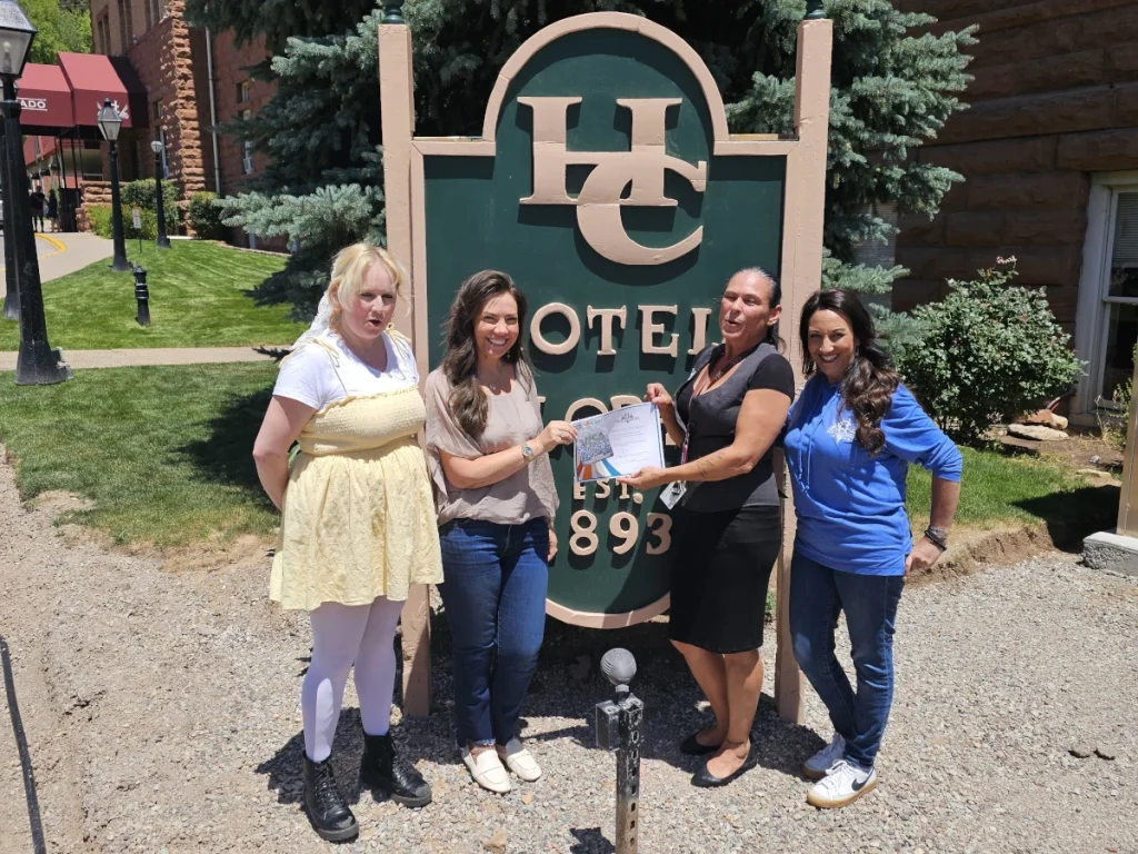 Four women stand outside in front of a green hotel sign, smiling at the camera. Two of them hold a certificate together, celebrating their achievement at the John Macdonald location. Trees, grass, and a building are visible in the background.