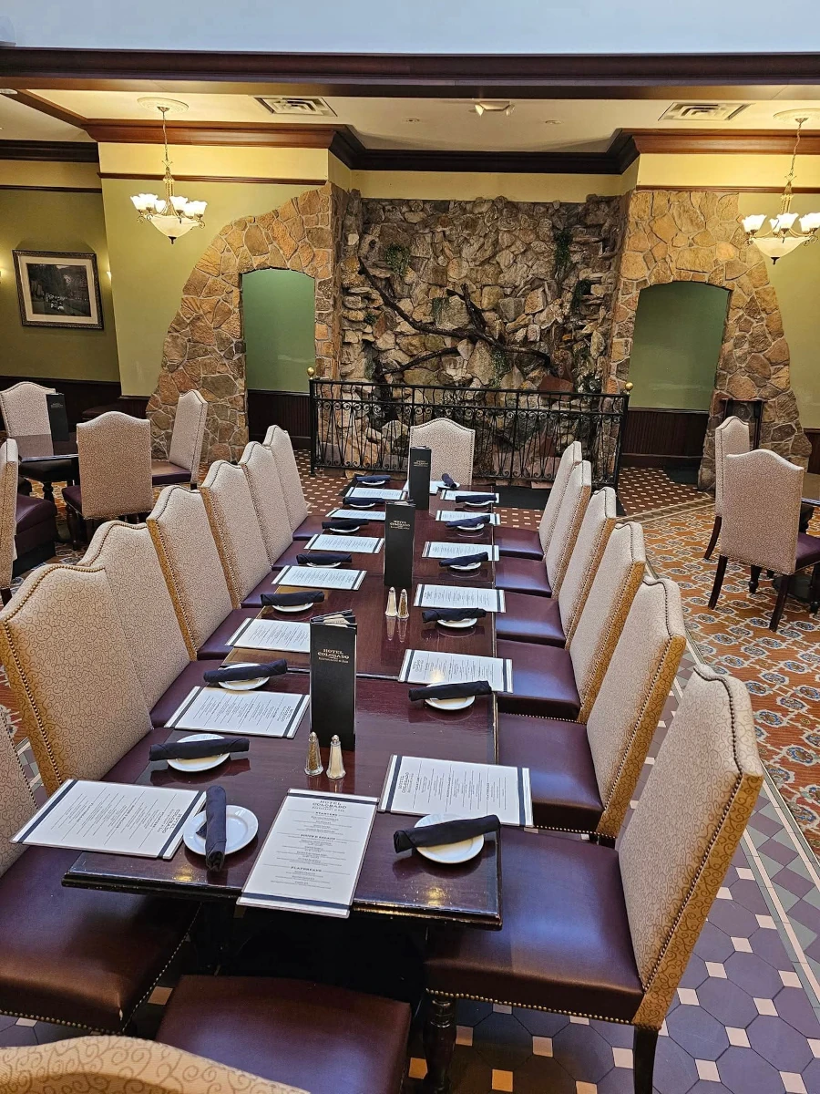 A long dining table set with menus, plates, and utensils is surrounded by upholstered chairs in an elegant restaurant, featuring tiled floors and a striking stone accent wall.