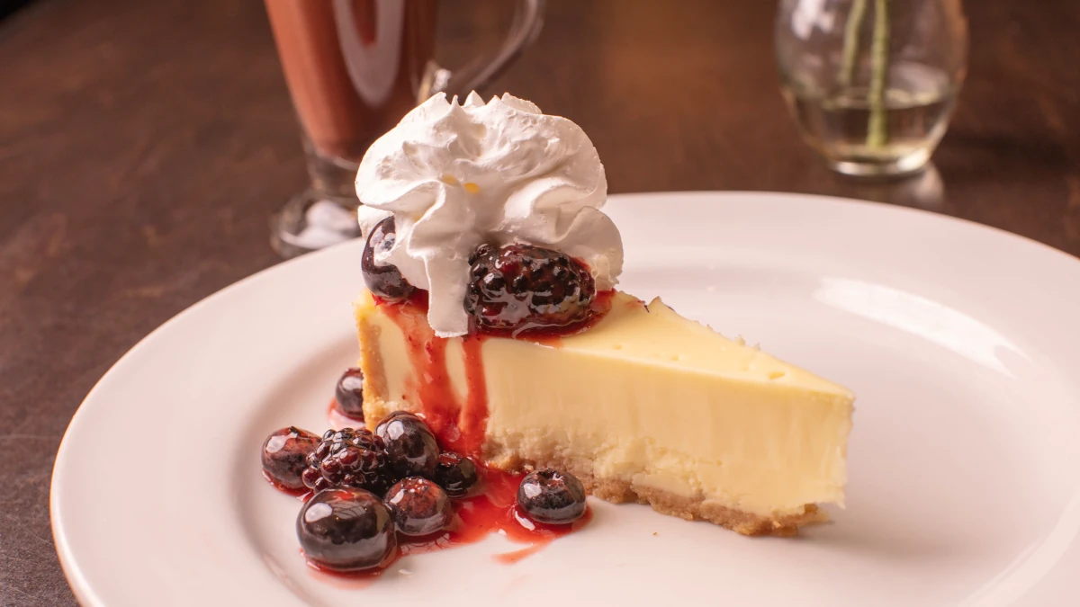 A slice of cheesecake topped with whipped cream and berry compote sits on a white plate at the restaurant, accompanied by a glass of chocolate drink and a vase in the blurred background.