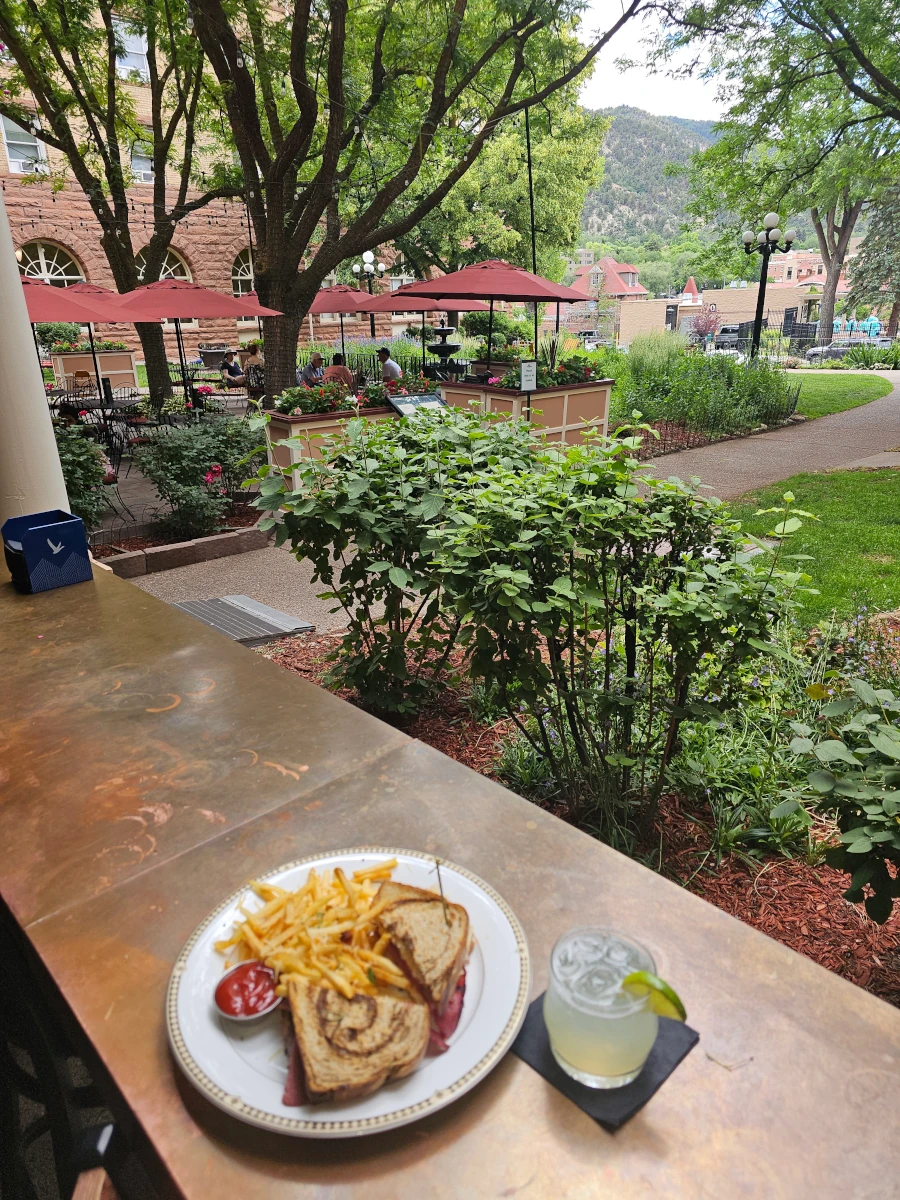 A plate with a sandwich, fries, and ketchup sits on a table at Coppertop, overlooking a garden patio with outdoor seating, trees, and distant buildings under a cloudy sky. A drink with lime rests on a black napkin beside the plate.