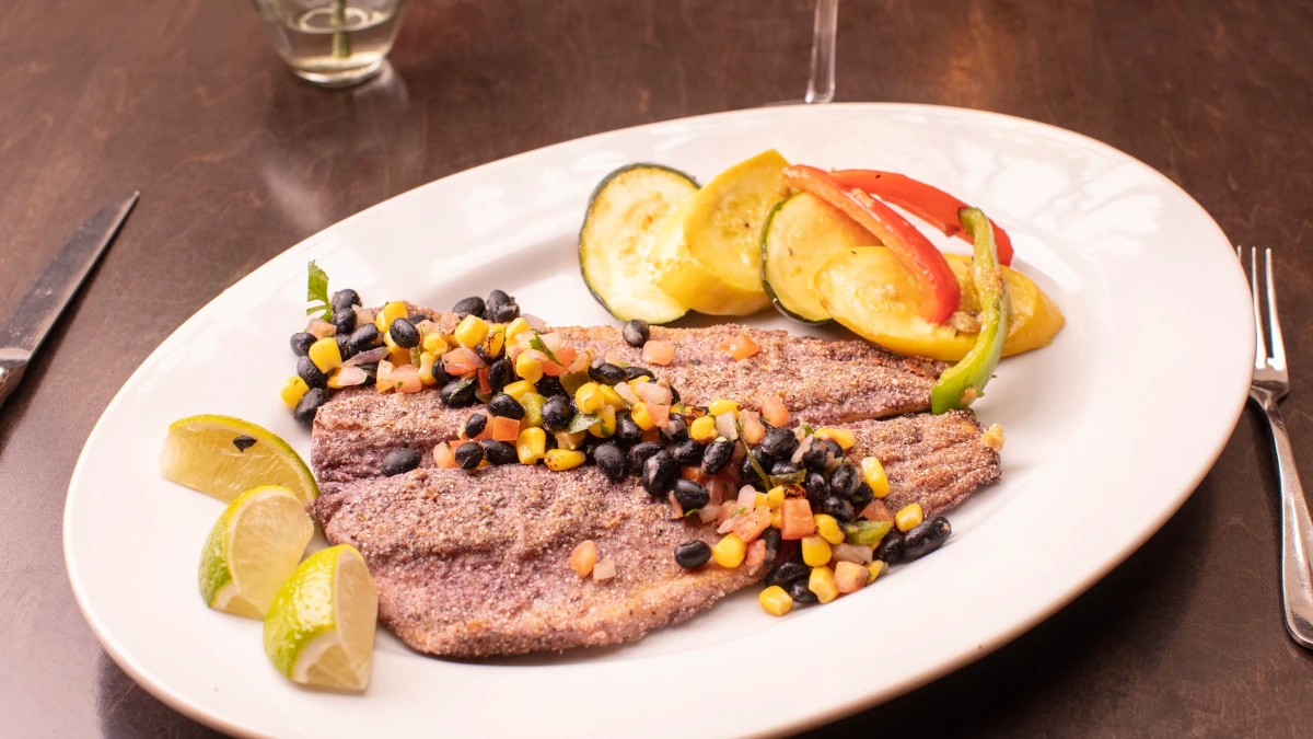 A white plate with two pieces of blackened fish topped with diced tomatoes, corn, and black beans is served at the restaurant with sautéed zucchini, yellow squash, red bell pepper, and lime wedges on a dark wooden table.
