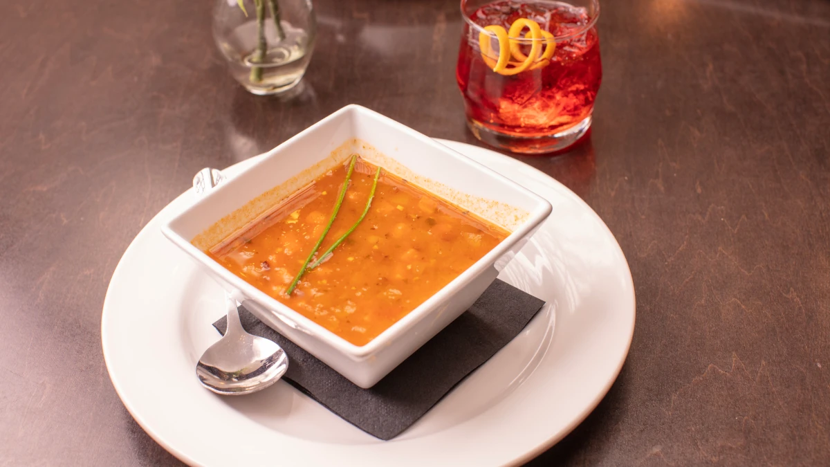A square white bowl of soup garnished with chives sits on a white plate with a spoon, next to a glass of red cocktail with orange peel, on a dark wooden restaurant table.