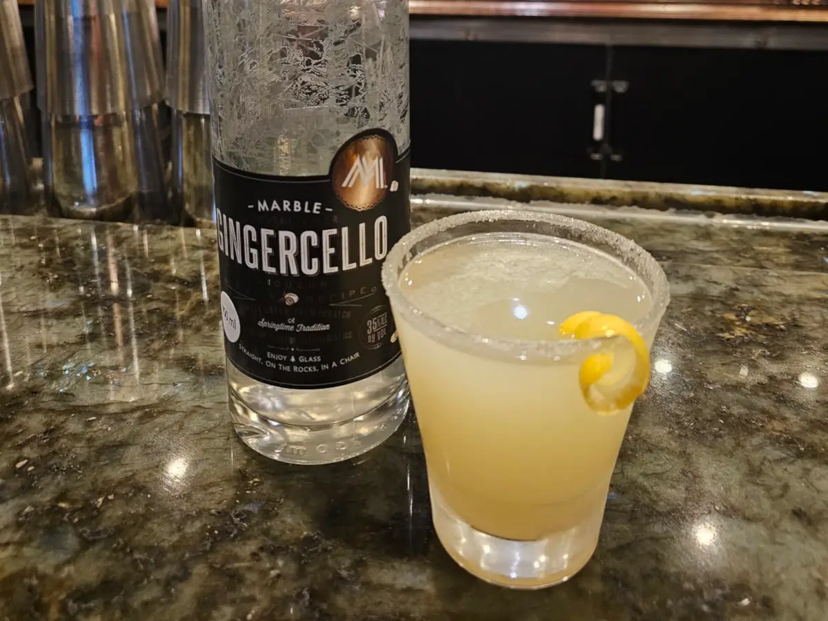 A glass of pale yellow Ginger Drop cocktail with a sugared rim and lemon twist garnish beside a bottle labeled Gingercello.