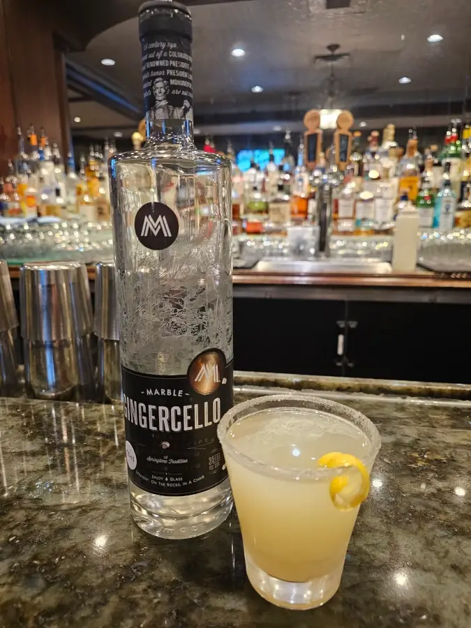 A bottle of Marble Gingercello liqueur stands beside a ginger drop cocktail garnished with a lemon twist on a marble bar counter.